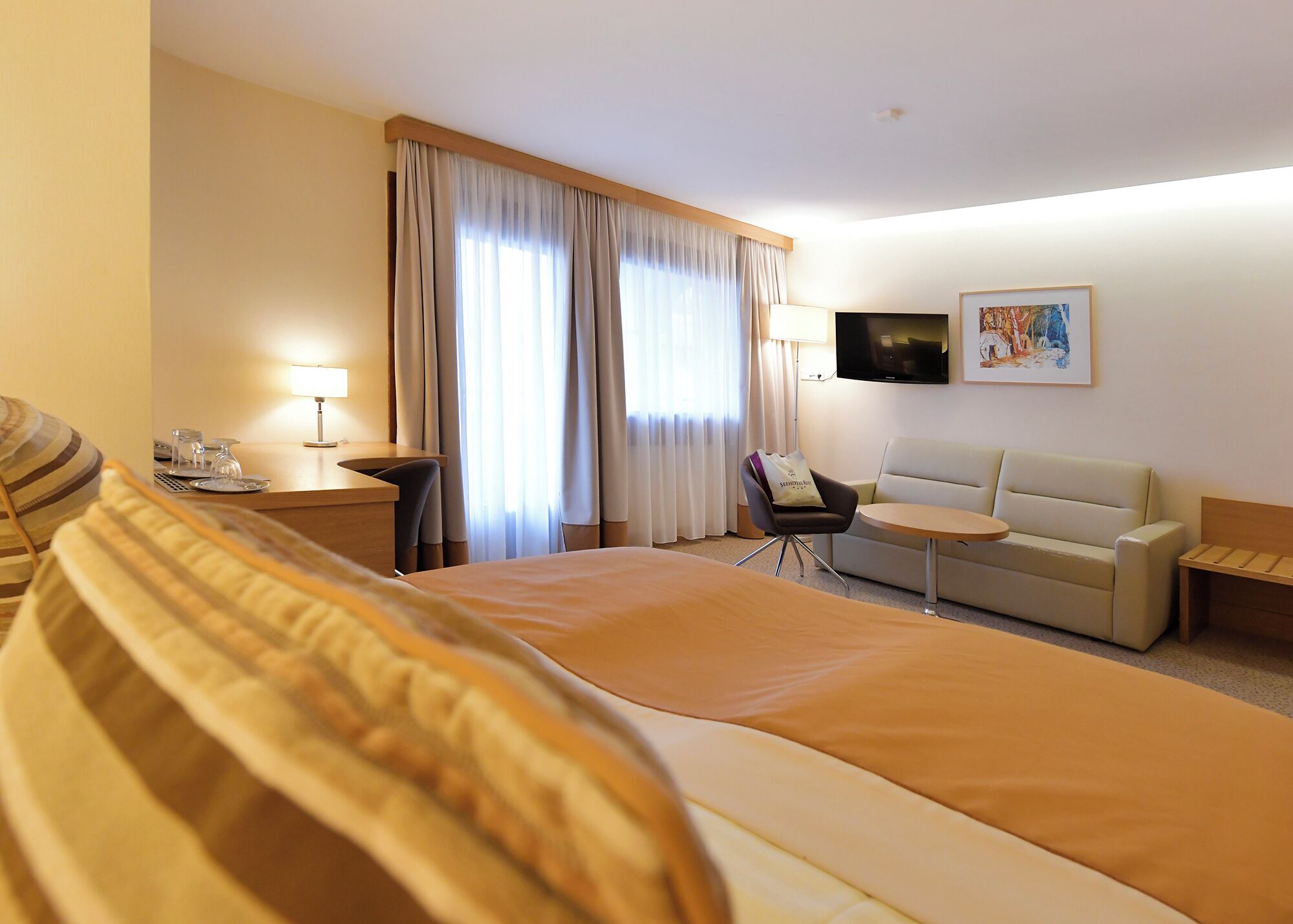 A hotel room with a large bed, a sofa, a small table, a chair, a TV, and a window with curtains.