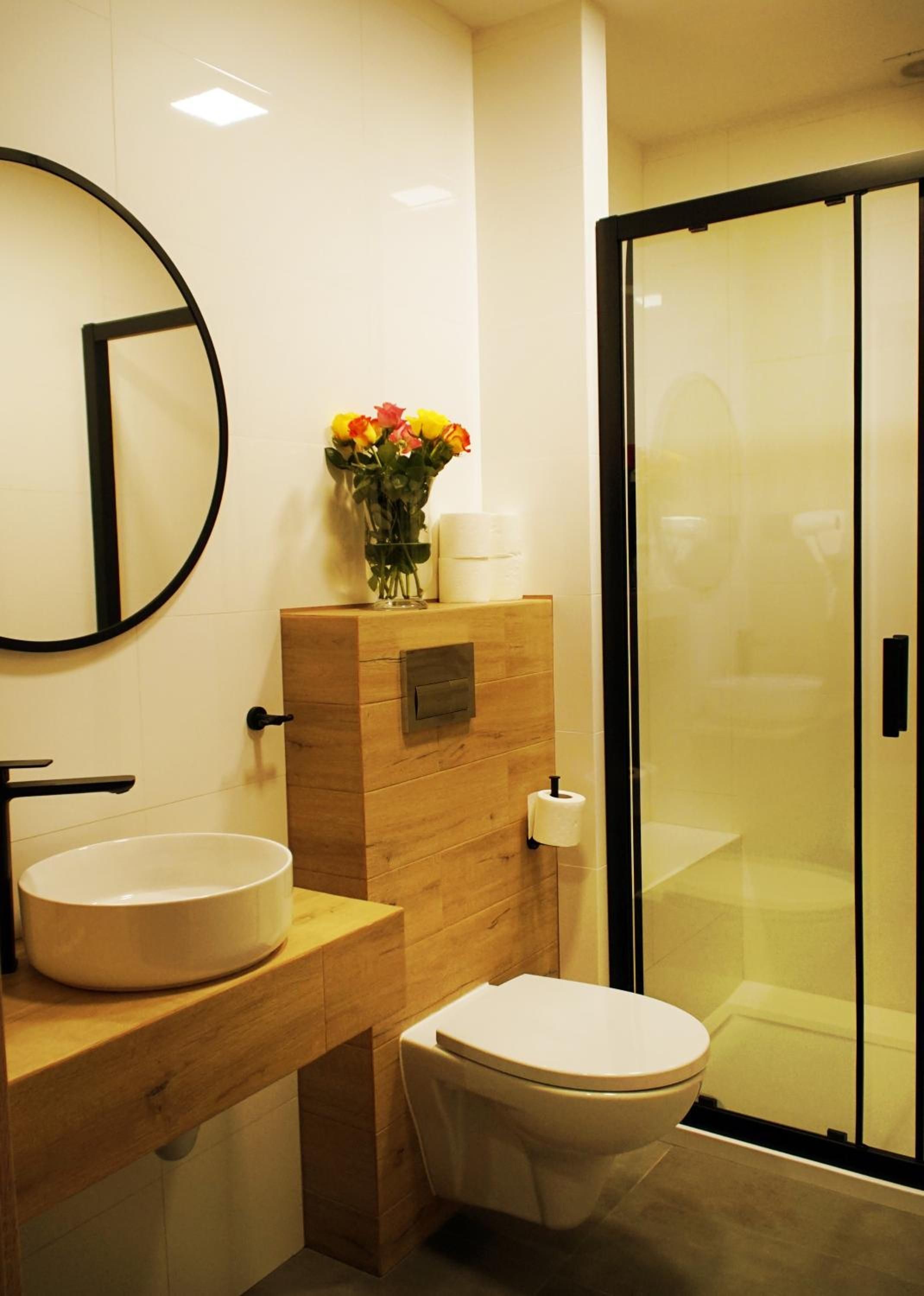 Ein modernes Badezimmer mit rundem Spiegel, einem Holzanrichte, einer Toilette und einer Duschkabine aus Glas.