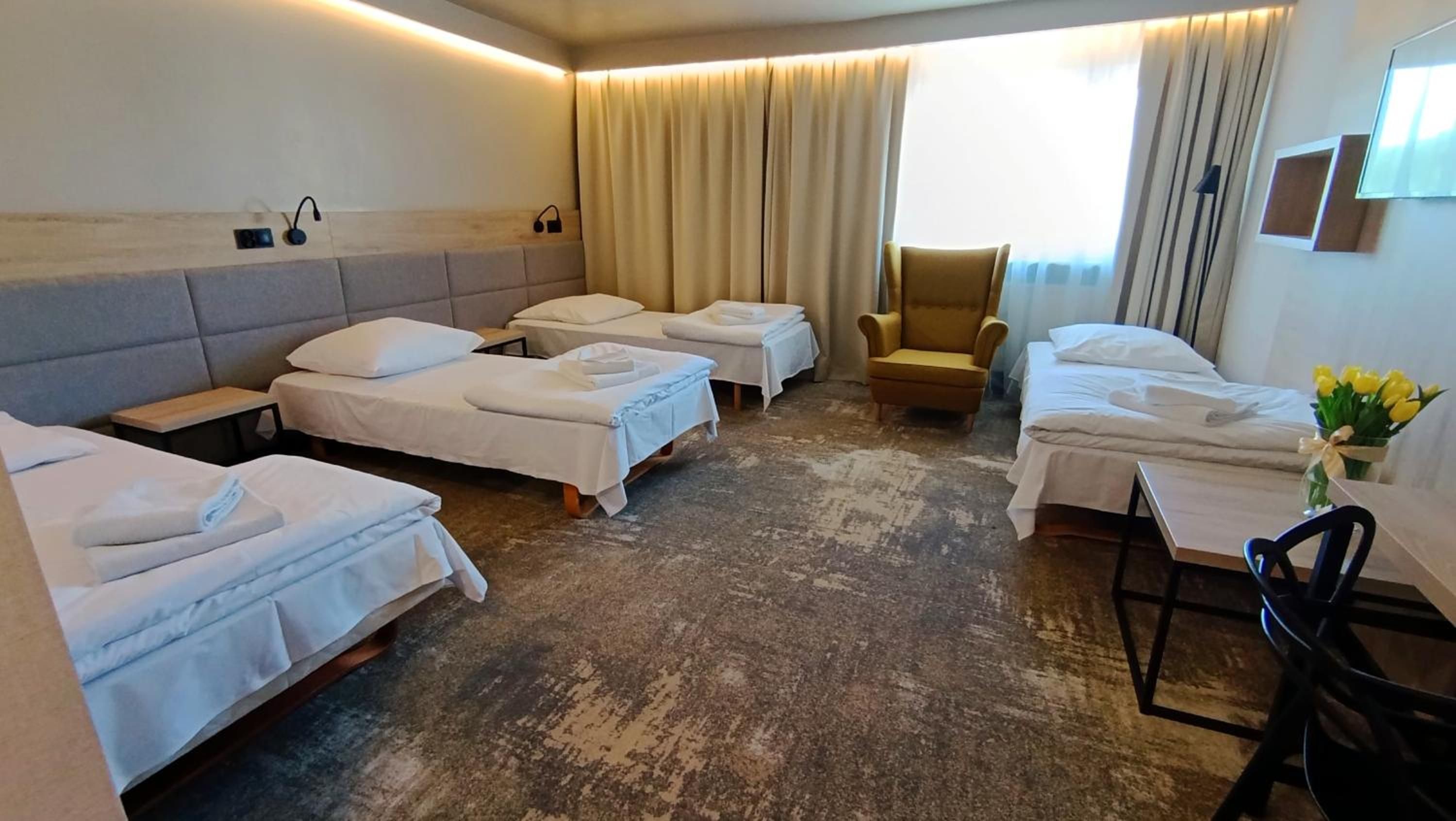 Habitación de hotel con cuatro camas individuales, una silla, un escritorio y un ventanal con cortinas.