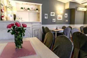 A dining area with a table set for four, chairs, a vase with flowers, a shelf with decorative items, and wall-mounted pictures. - Hotel Relax (Kety)