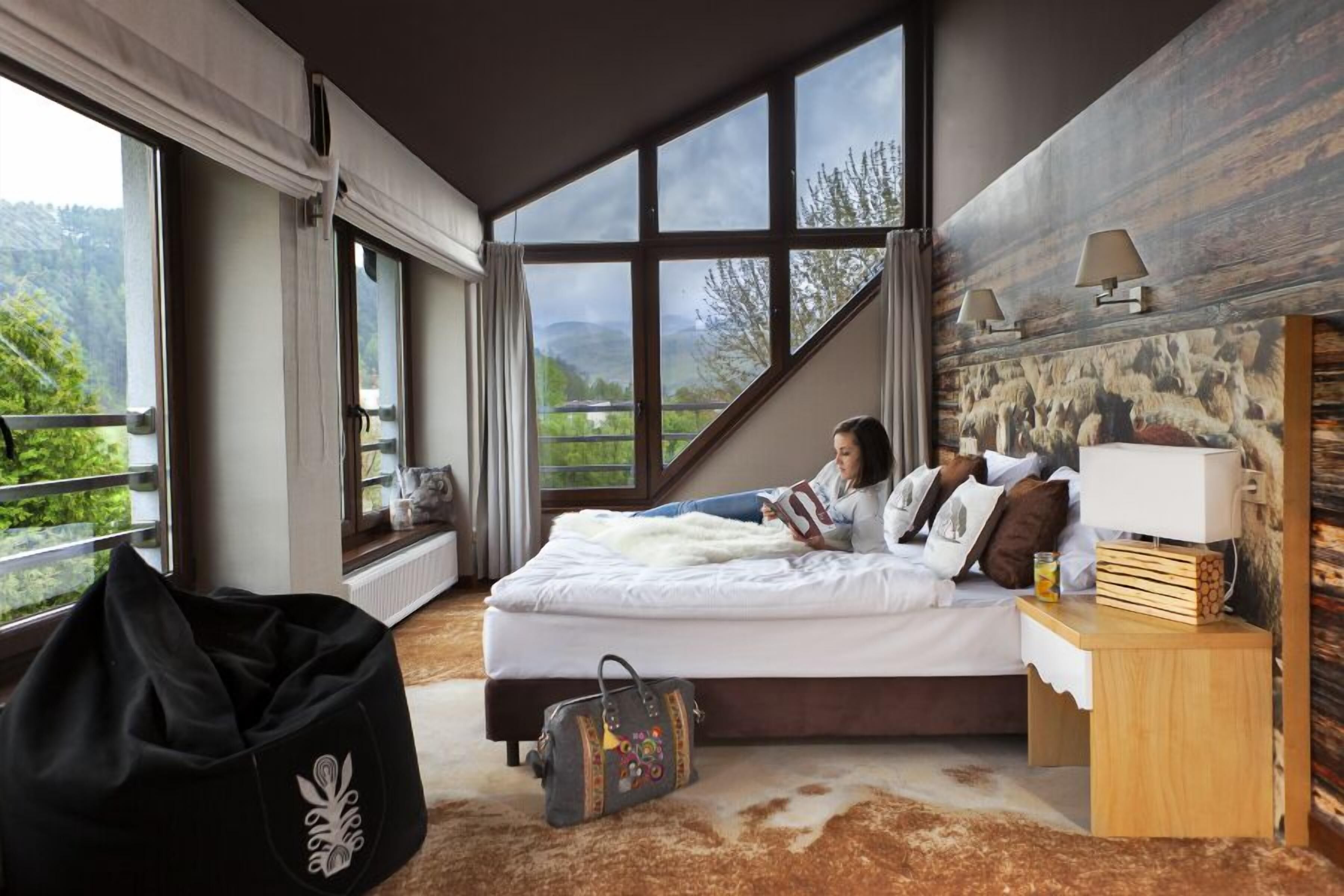 A bedroom with a large window, a bed, a bedside table with a lamp, and a view of greenery outside.