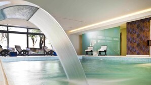 Una piscina interior con cascada, sillones de descanso y vistas a árboles fuera.