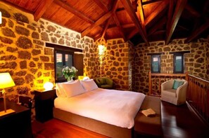 A bedroom with a stone wall, wooden ceiling beams, a bed, a chair, and a window.