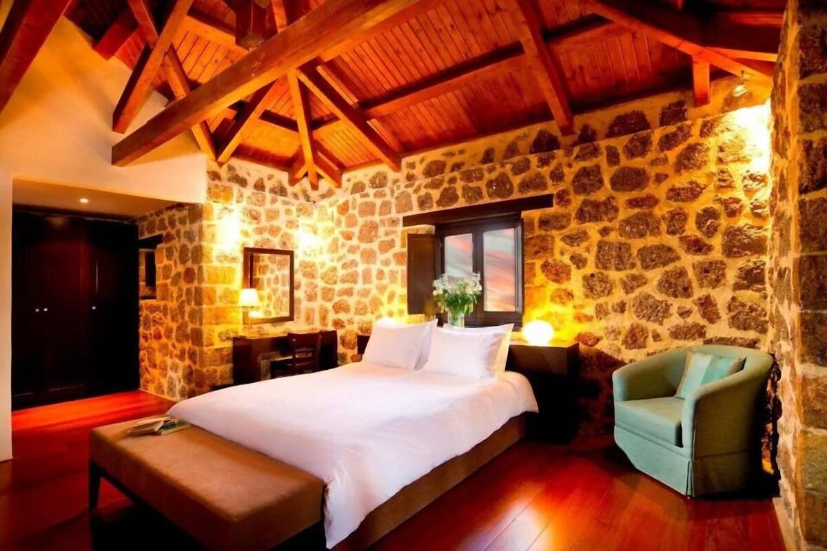 A cozy room with a stone wall, wooden ceiling beams, a bed, a chair, and a nightstand with a lamp.