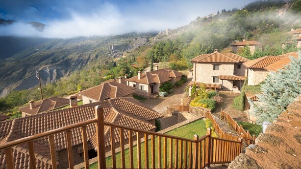 Un pueblo en la ladera de una colina con casas de tejados de terracota, una escalera de madera y vistas de las montañas circundantes.