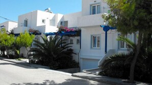 A row of white buildings with blue accents, likely residential, with a clear sky and some greenery. - Atlantis Place Milos (Milos)