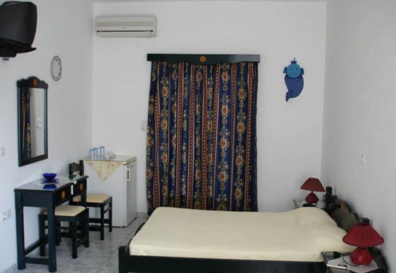 Una habitación pequeña y sencilla con una cama, un escritorio, una silla, una mesita, una lámpara roja y una cortina con estampado.