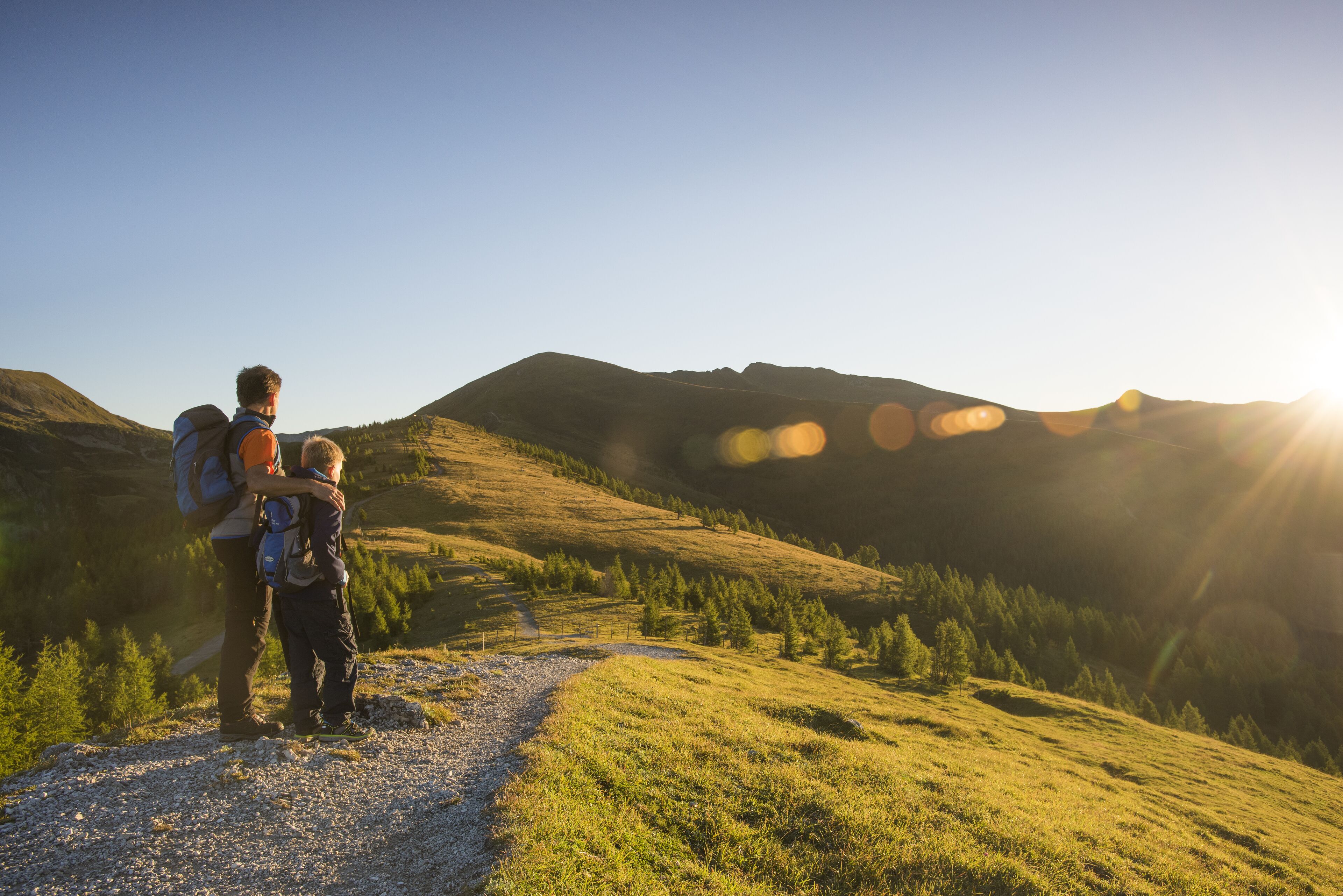 A person with a backpack and a child on a gravel path in a hilly area with trees and mountains in the background.