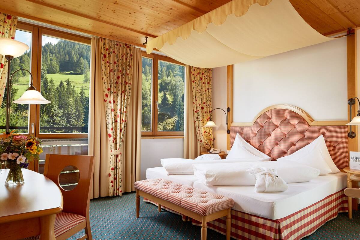 A hotel room with a bed, a bench, a table with a vase of flowers, and curtains with a floral pattern.