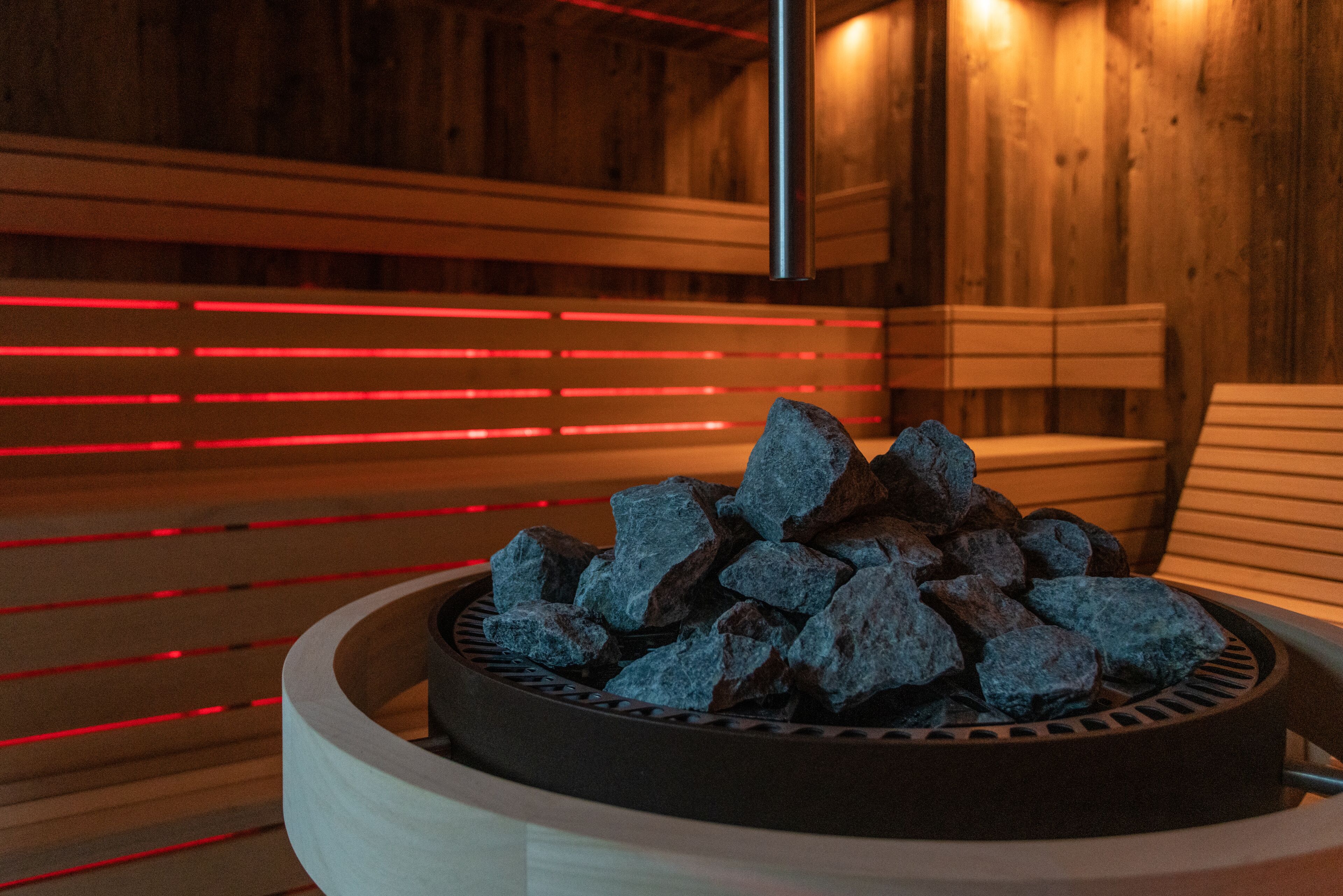 A sauna with a circular seating area filled with rocks, wooden benches, and red ambient lighting.