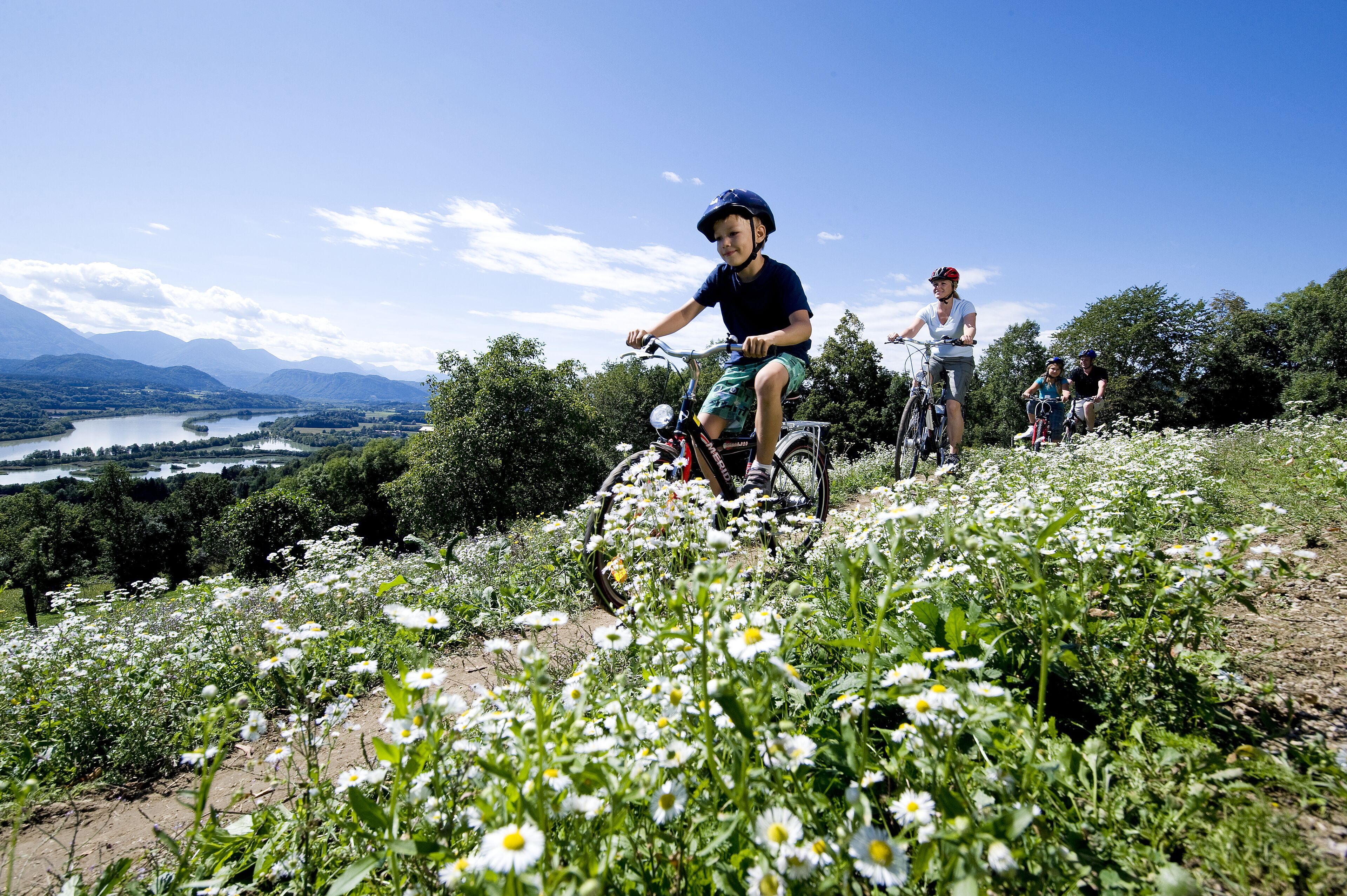 Un groupe de personnes fait du vélo sur un chemin de terre, entouré de fleurs blanches et d’arbres verts. En arrière-plan, on aperçoit un paysage magnifique avec des montagnes et un lac.