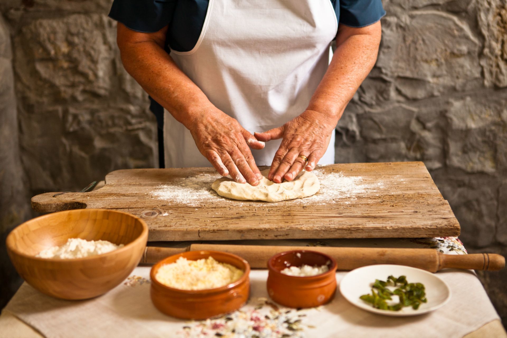 A person kneading dough on a wooden surface with bowls of ingredients nearby.