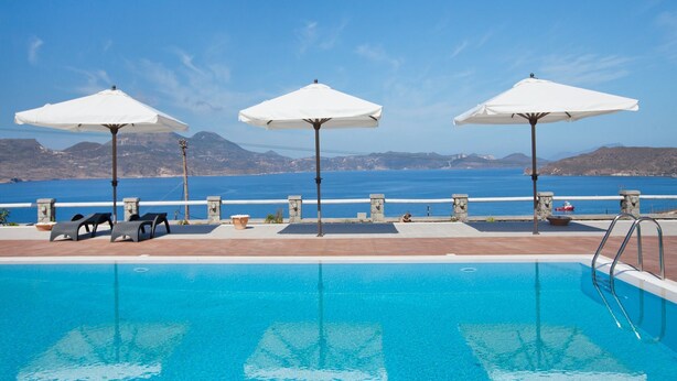 A swimming pool with clear blue water, surrounded by lounge chairs and umbrellas, overlooking a scenic view of the sea and mountains. - Miland Suites (Milos)