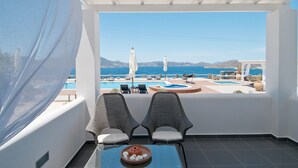 A balcony with two wicker chairs, a small table with a bowl of fruits, and a view of a pool and mountains. - Miland Suites (Milos)