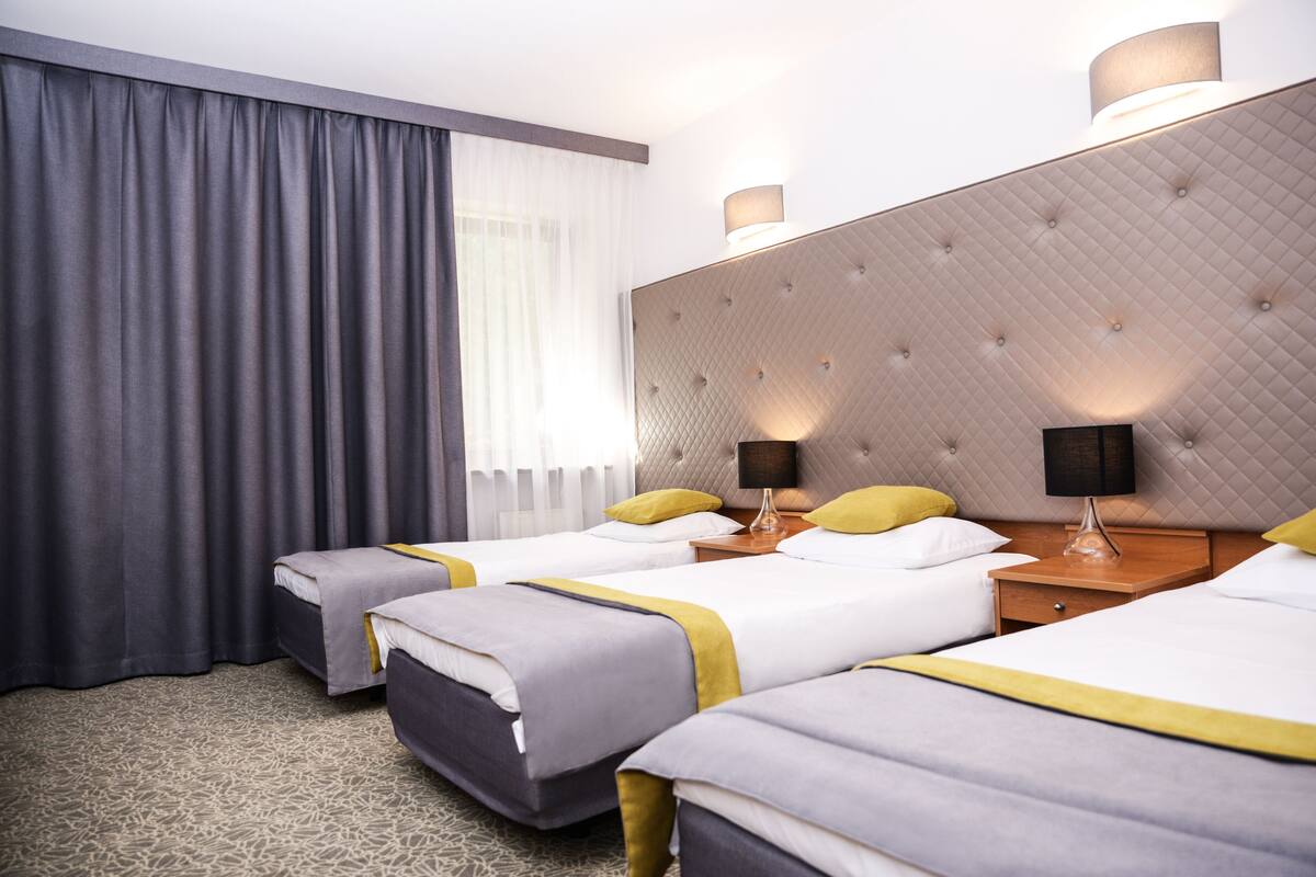 Hotellihuone, jossa on kolme sänkyä, suuri ikkuna verhoilla, koristeltu pääty ja yöpöytävalaisimet.