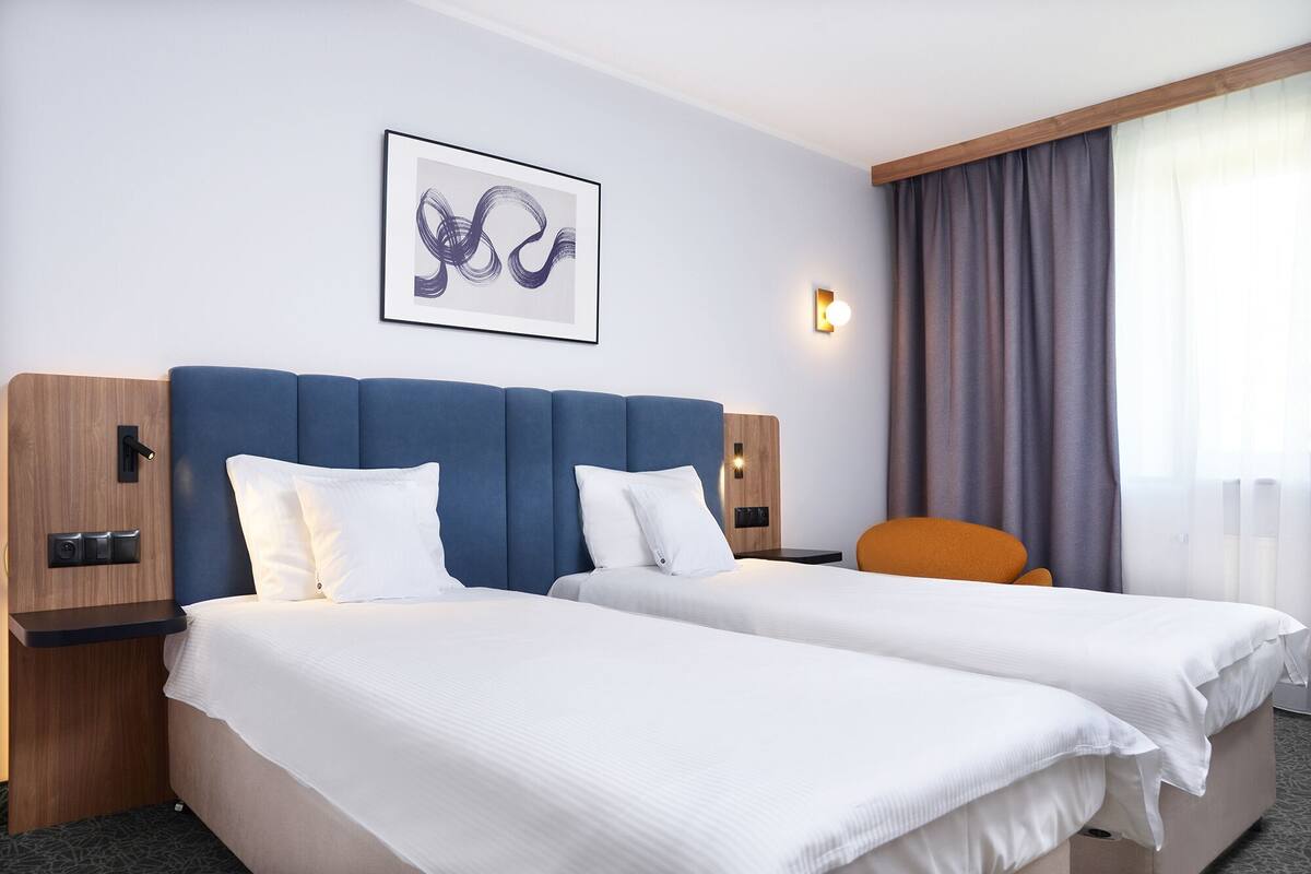 Hotellihuone, jossa on kaksi sänkyä, sininen pääty ja seinälle kiinnitetty taideteos sekä ikkuna, jossa on verhot.