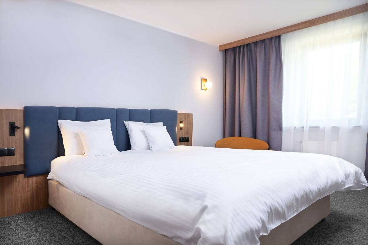 Hotellihuone, jossa on suuri sänky, puinen pääty, yöpöytä, seinään kiinnitetty lamppu ja ikkuna, jossa on verhot.