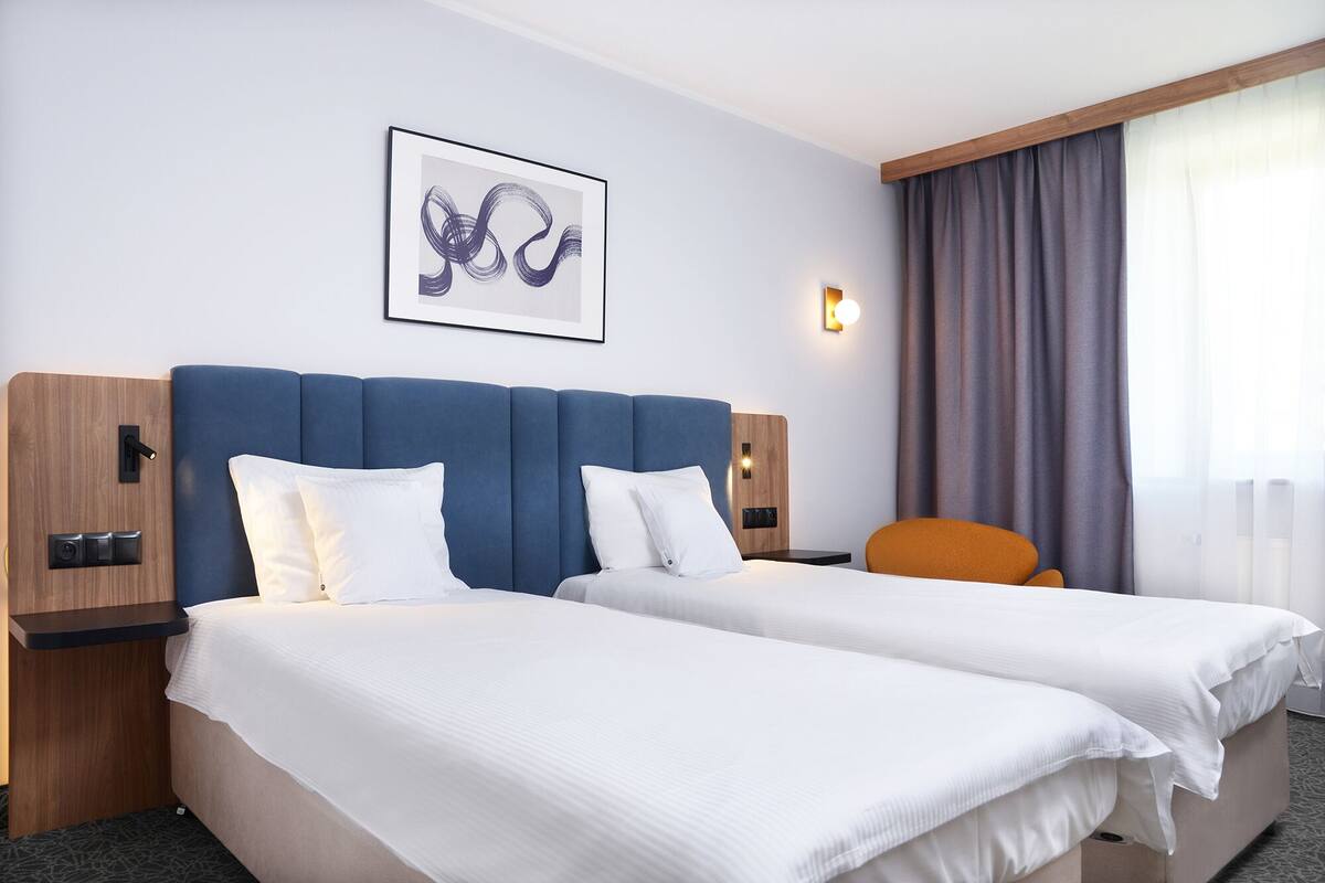 Hotellihuone, jossa on kaksi sänkyä, sininen pääty, puinen yöpöytä, seinään kiinnitetty valaisin ja ikkuna, jossa on verhot.