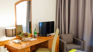 A hotel room with a wooden desk, a television, a chair, a mirror, and a window with curtains.