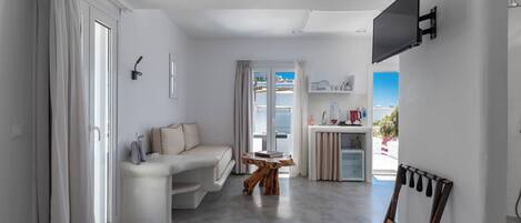 A modern hotel room with a white sofa, a wooden coffee table, a flat-screen TV mounted on the wall, and a view of the outdoors through the window.
