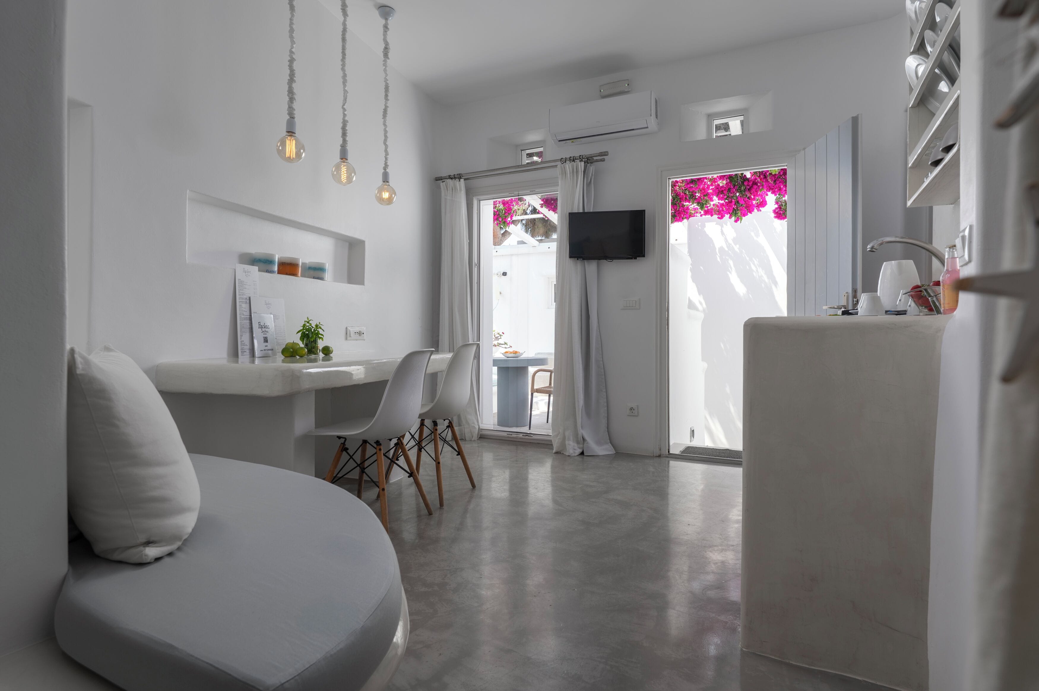 A modern, minimalist living room with a sofa, a dining area with chairs, a kitchen area with a fridge, and a TV mounted on the wall.