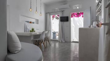 Uma sala de estar moderna e minimalista, com um sofá, uma área de refeições com cadeiras, uma cozinha com geladeira e uma TV fixada na parede.