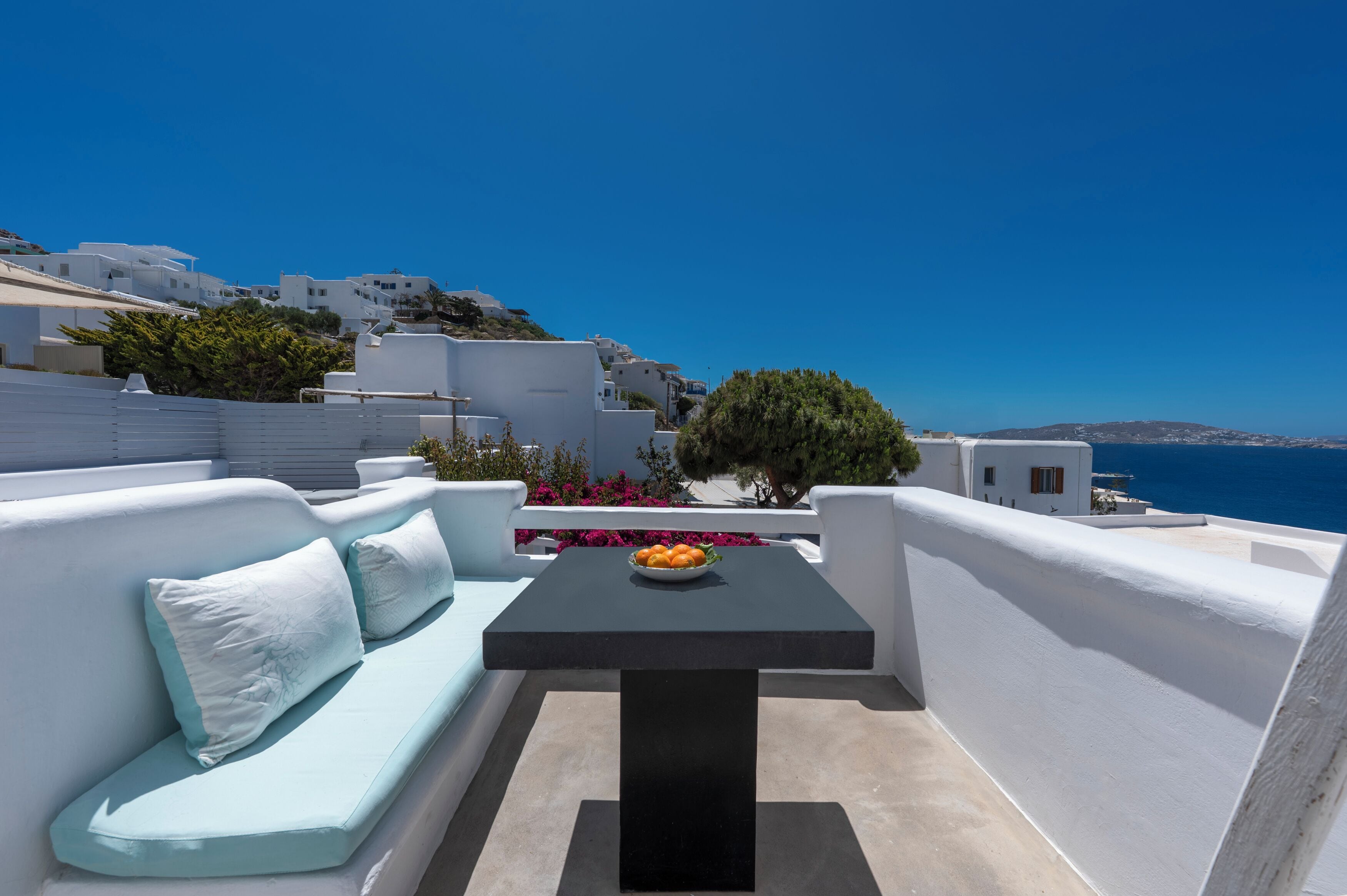 A balcony with a black table and a white sofa with light blue cushions, overlooking a coastal town with white buildings and a clear blue sky.