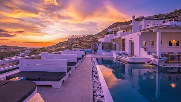 A poolside area with lounge chairs, a pool, and a view of a hillside with buildings. - Flaskos Luxury Suites (Mykonos)
