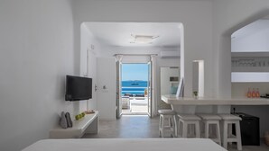 A modern hotel room with a TV, a bench, a small table with bottles, and a view of the sea. - Flaskos Luxury Suites (Mykonos)