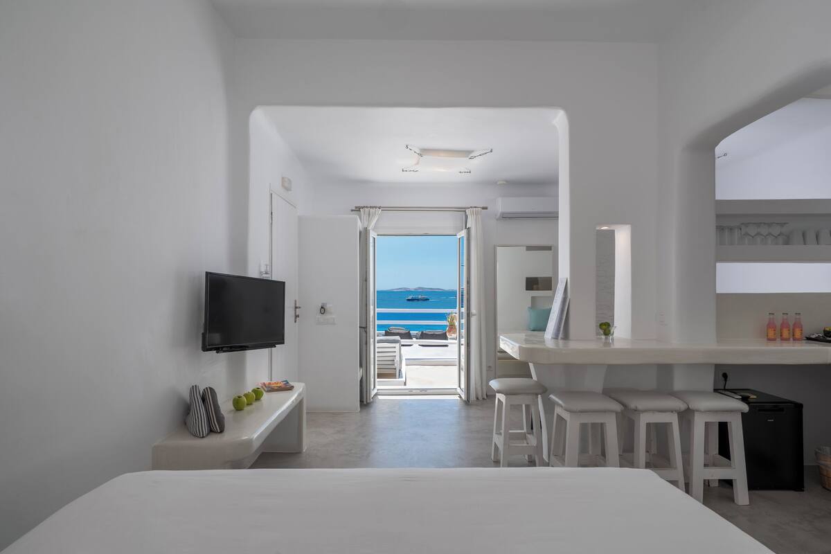 A modern hotel room with a TV, a bench, a small table with bottles, and a view of the sea.