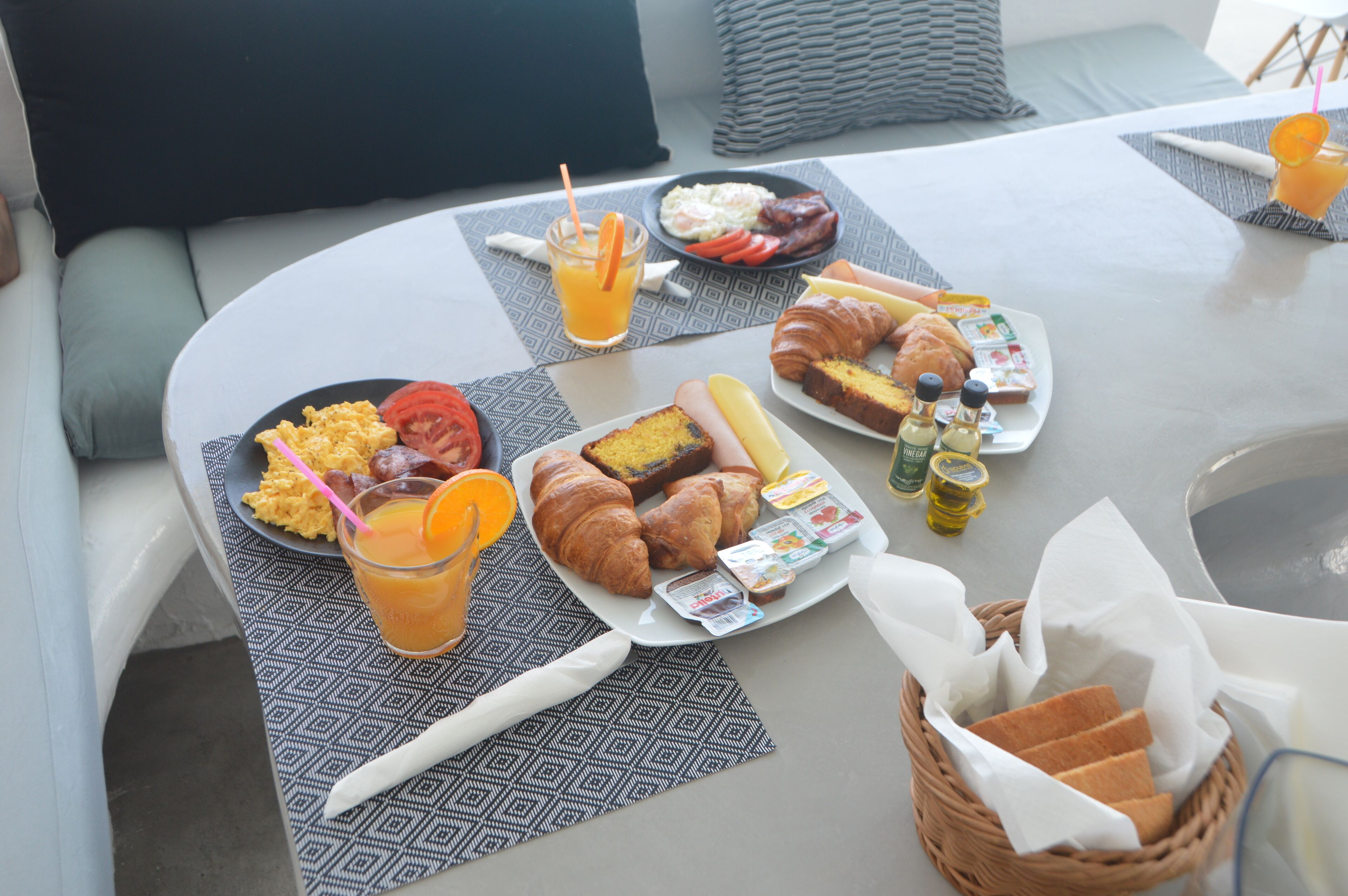 Une table de petit-déjeuner avec des assiettes remplies de nourriture, un panier de pain et un verre de jus d’orange.