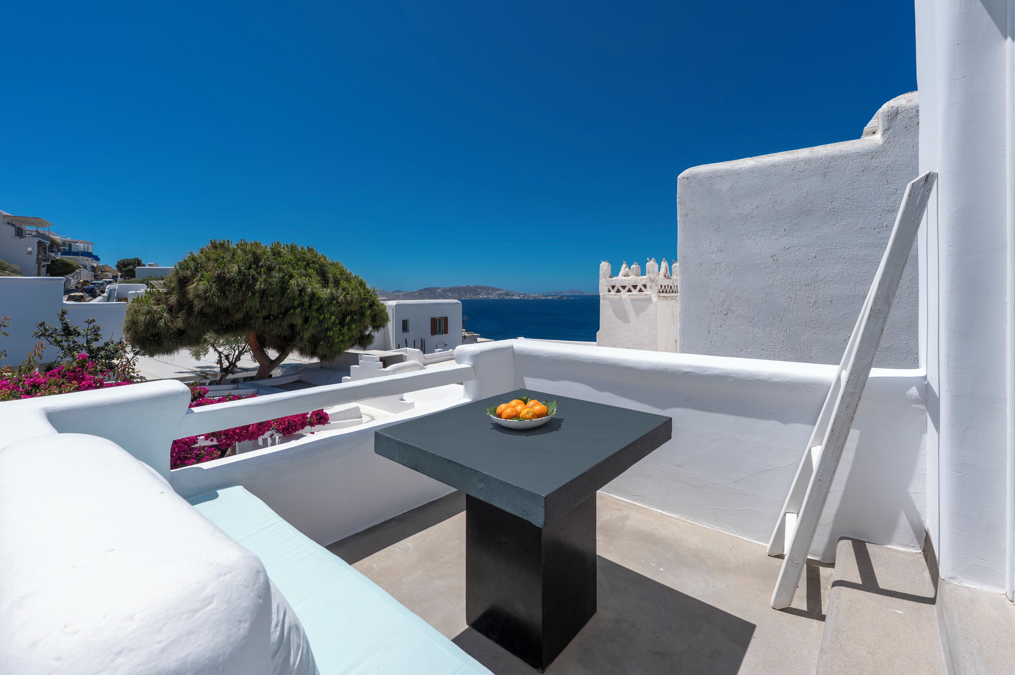 Una terraza de paredes blancas con una mesa, sillas, un árbol y vista al mar.