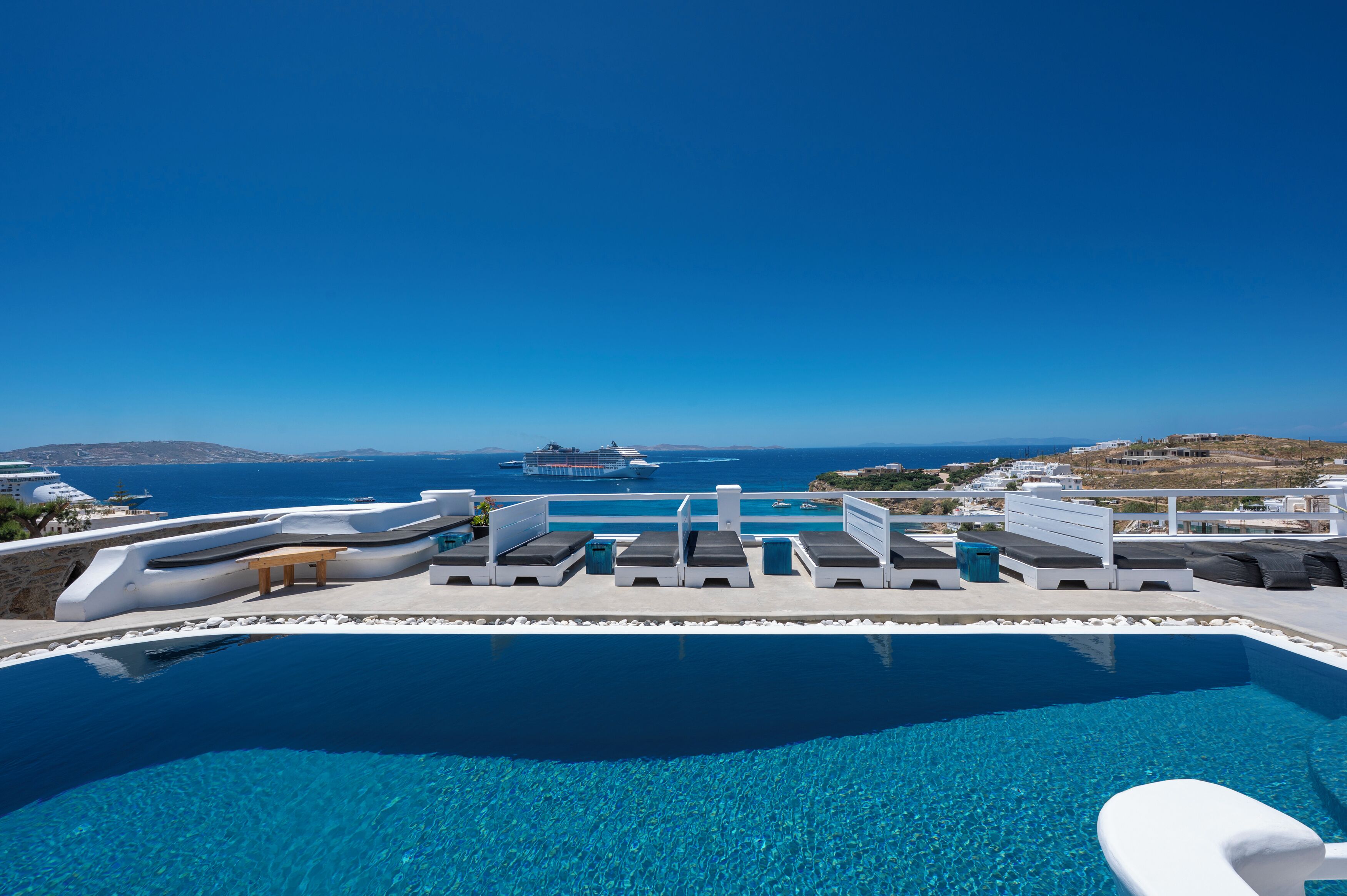 Un cielo azul despejado, una piscina con tumbonas y vistas al mar y edificios lejanos.