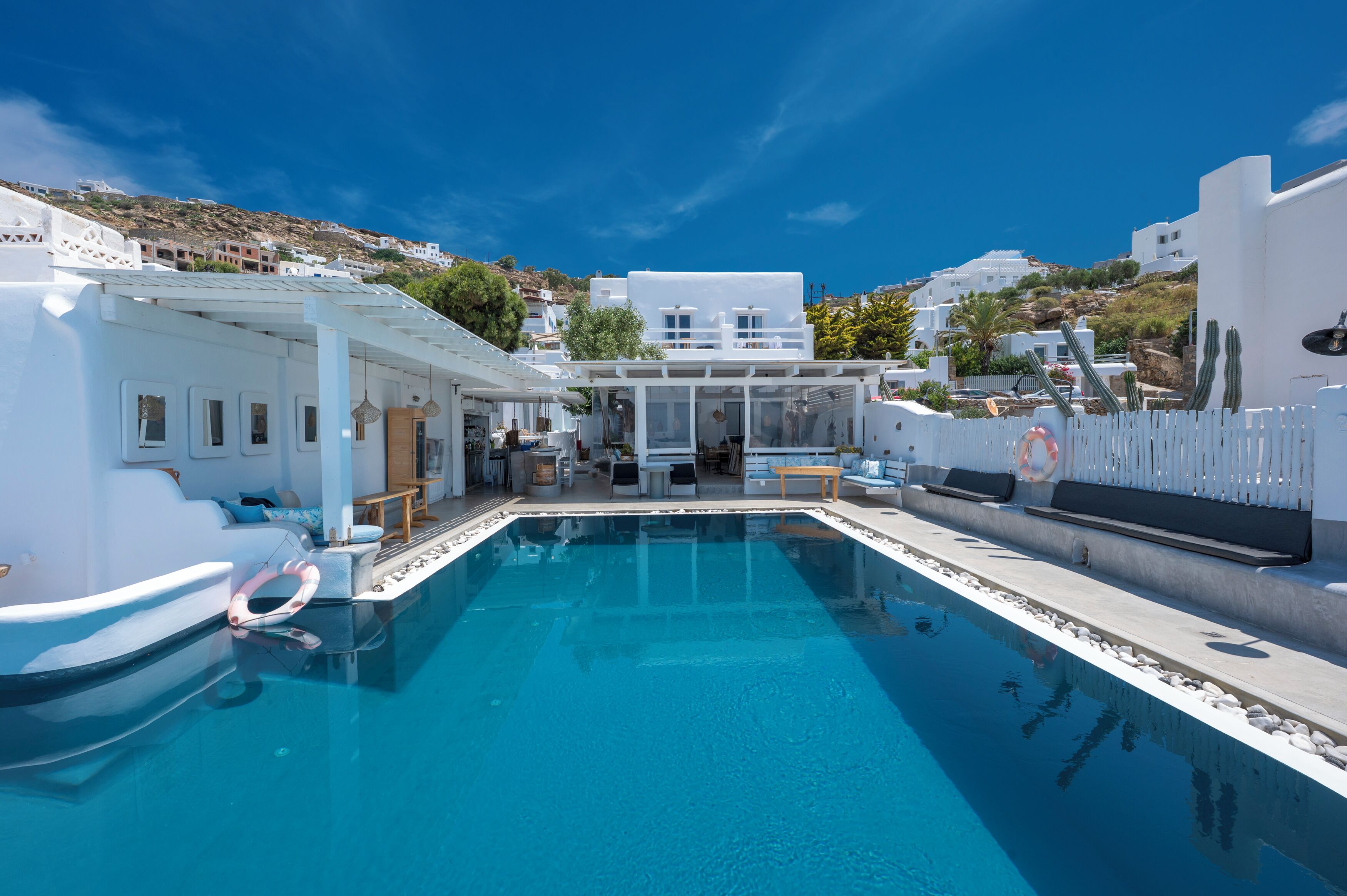 A modern pool area with a clear blue pool, white buildings in the background, and a white fence surrounding the pool.