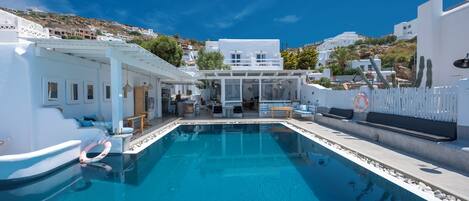 A modern pool area with a clear blue pool, white buildings in the background, and a white fence surrounding the pool.