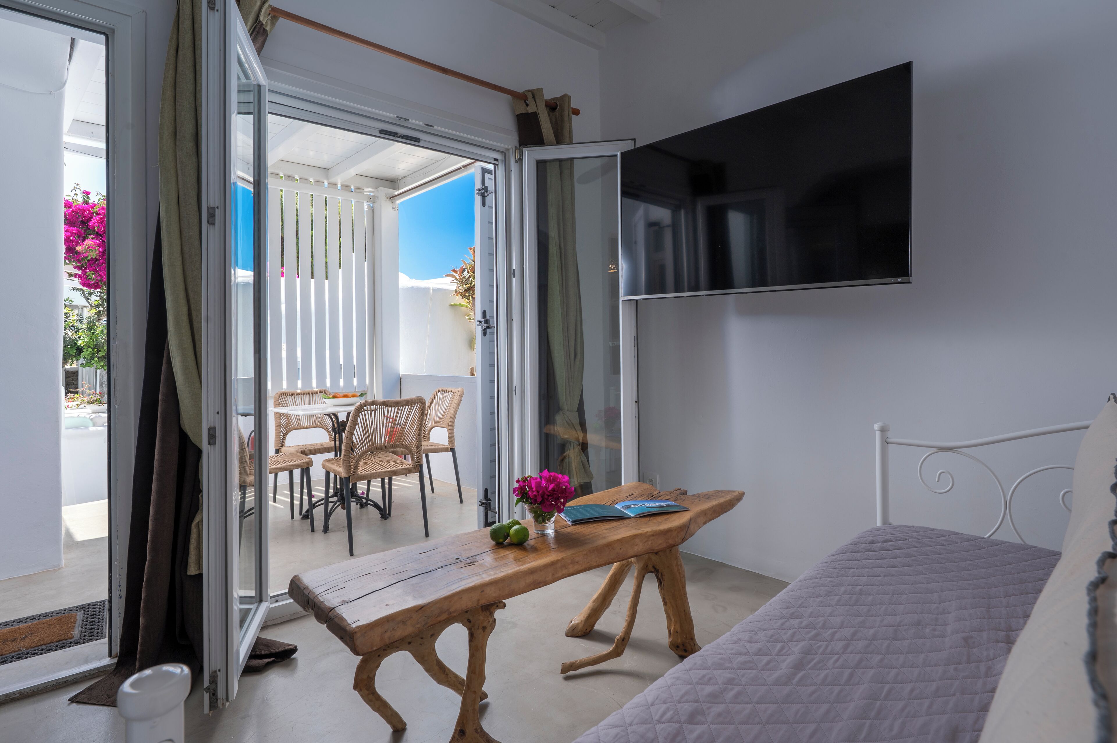 Un dormitorio con una cama, una mesa de madera con un jarrón de flores, un televisor montado en la pared y una puerta corrediza de vidrio que da a un balcón con mobiliario exterior.