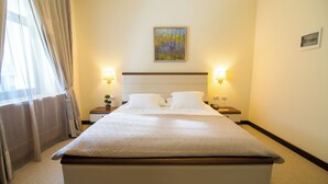 A hotel room with a large bed, two bedside tables, a nightstand, a painting on the wall, and a window with curtains. - Kotoni (Tirana)