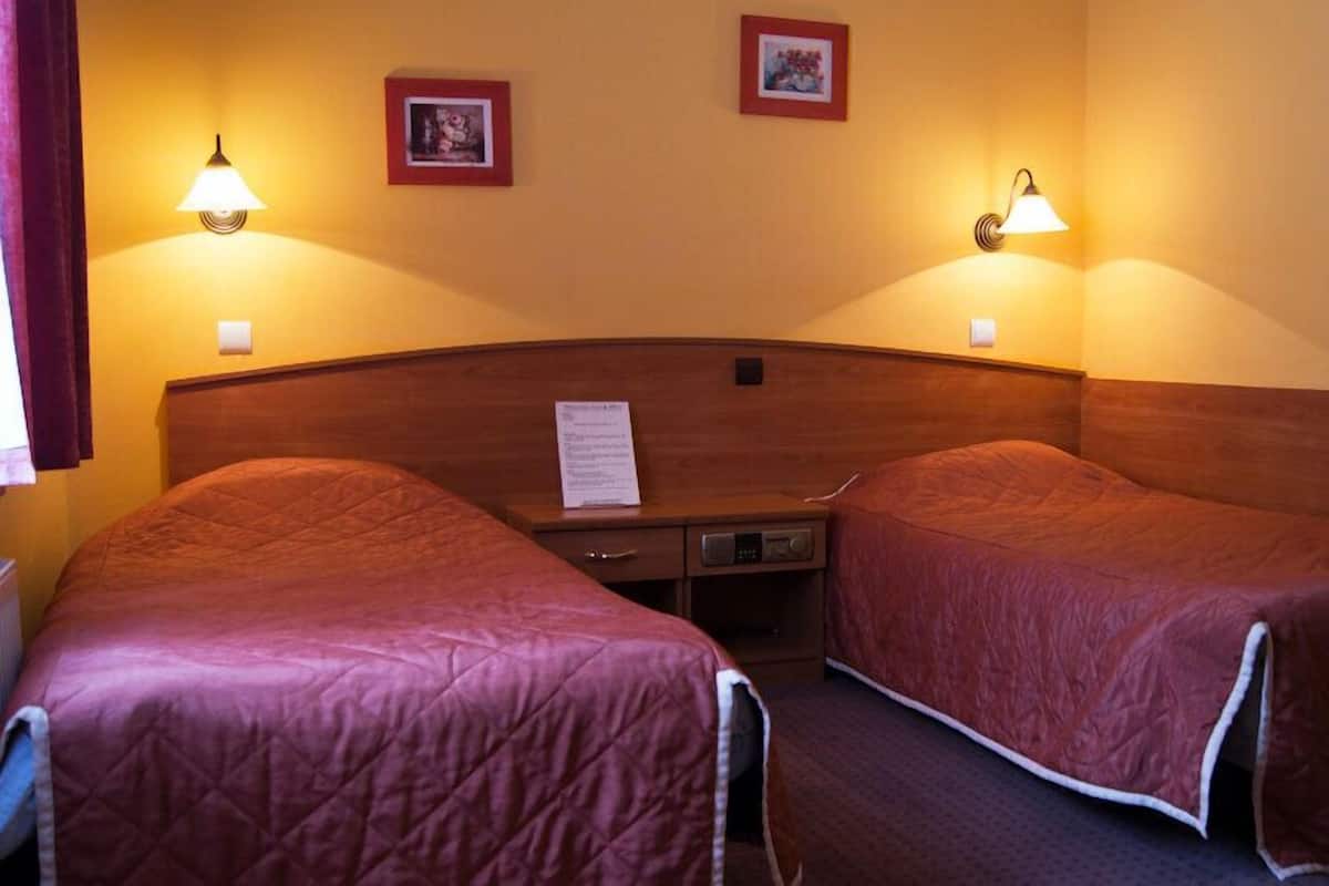 Habitación de hotel con dos camas individuales, cabeceros de madera, lámparas de noche y una mesita con lámpara.