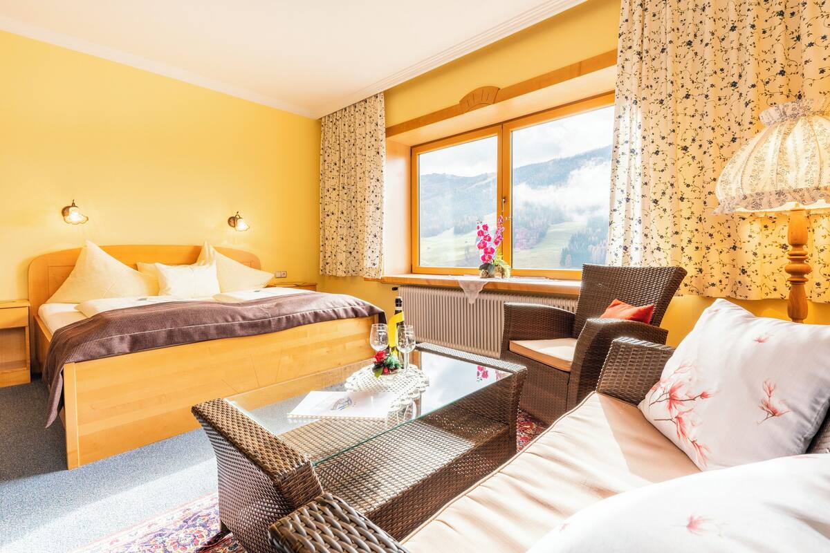 A hotel room with a bed, a sofa, a small table with a bottle and glasses, and a view of mountains through a window.
