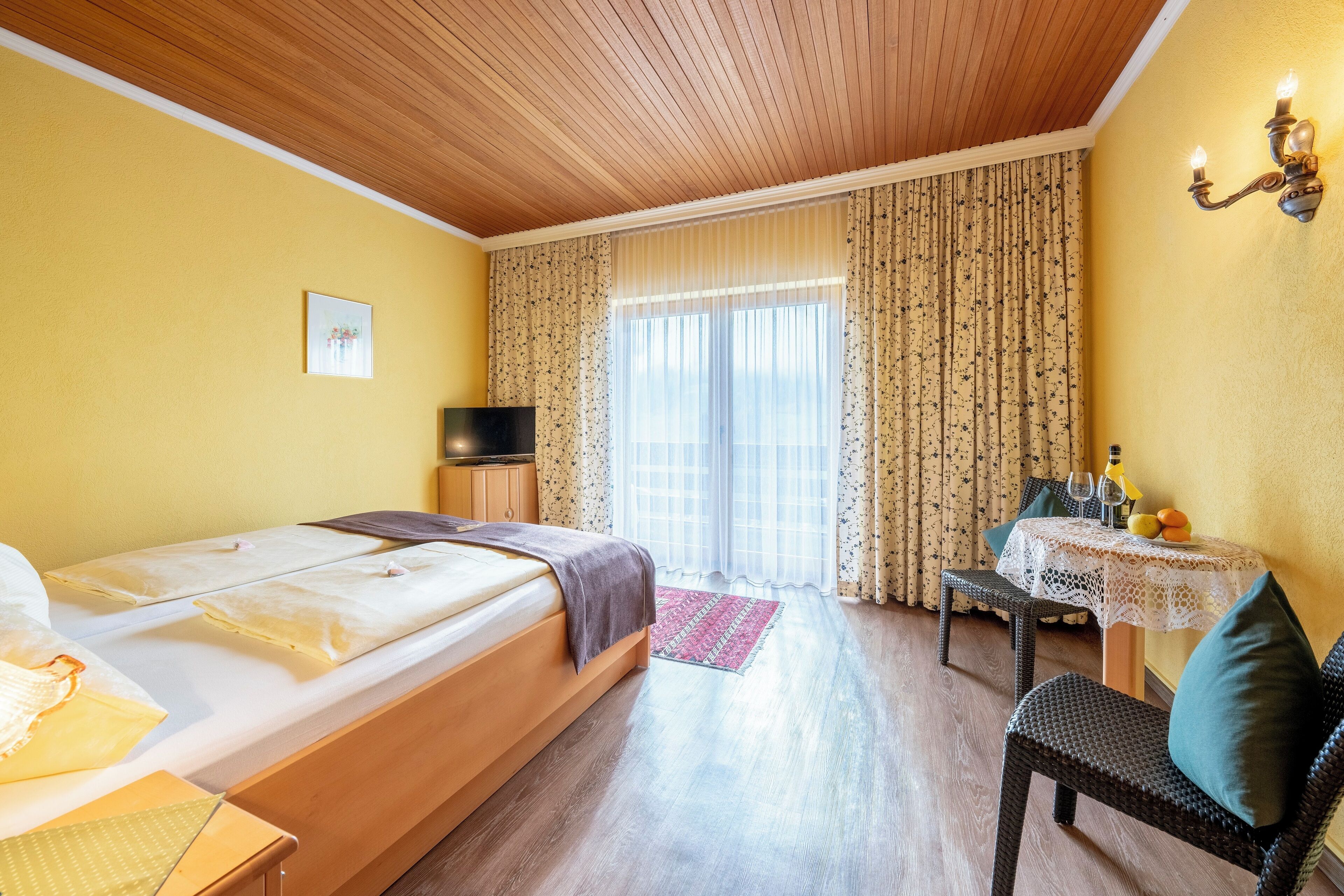 A hotel room with a wooden ceiling, a bed with a TV, a dining table with a lamp, and a window with curtains.