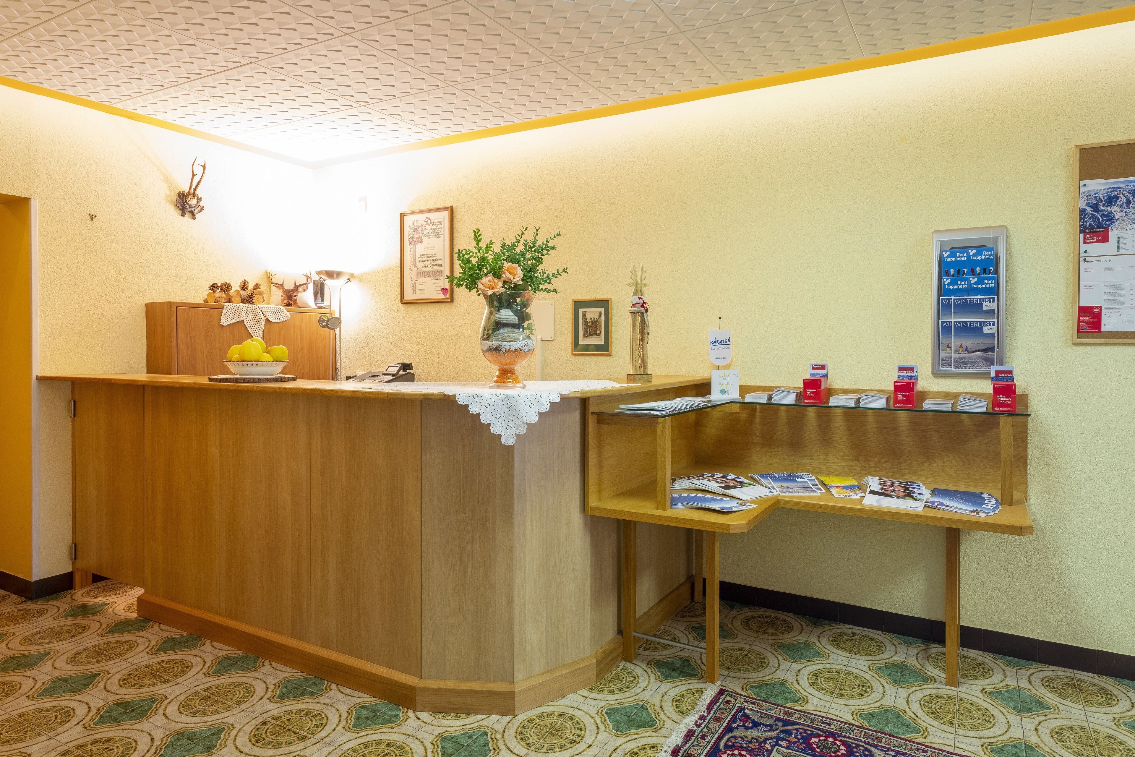 A reception area with a wooden counter, a vase with flowers, a bulletin board with notices, and a patterned floor.