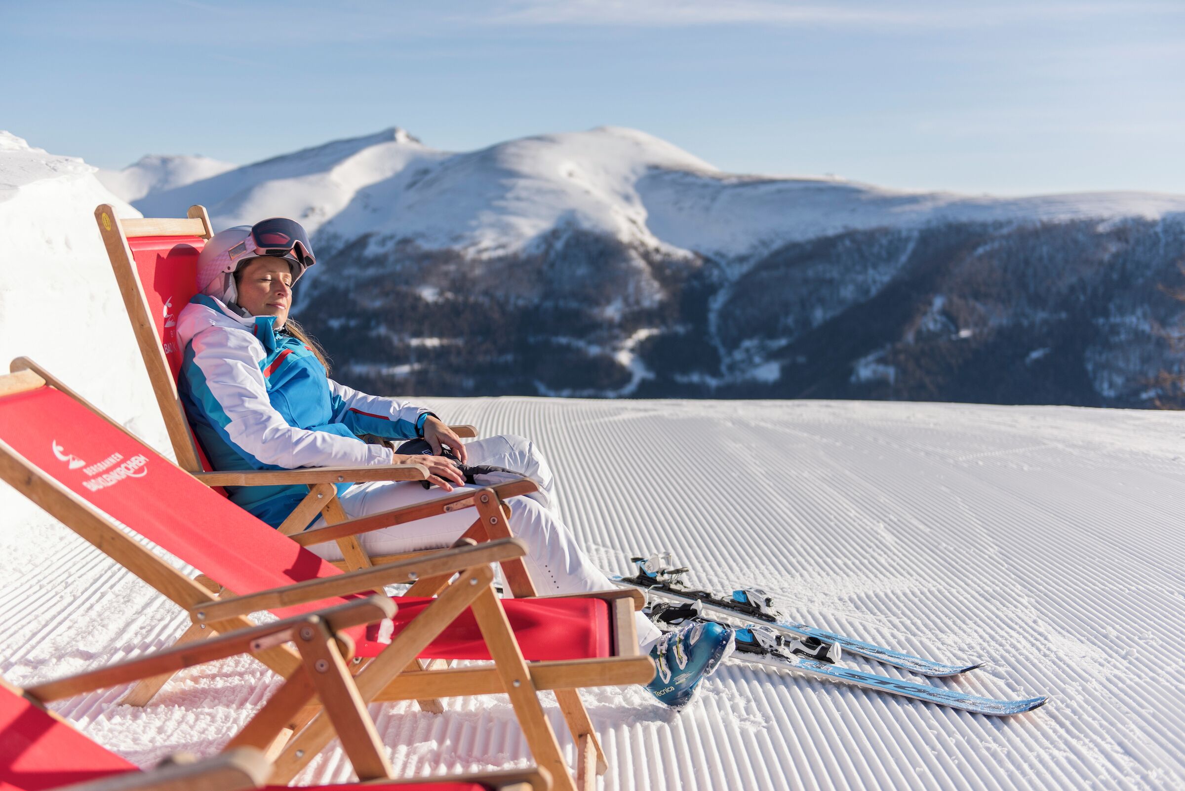 Una persona sentada en una silla reclinable roja, con esquíes y bastones de esquí a su lado, en una pendiente nevada con montañas al fondo.