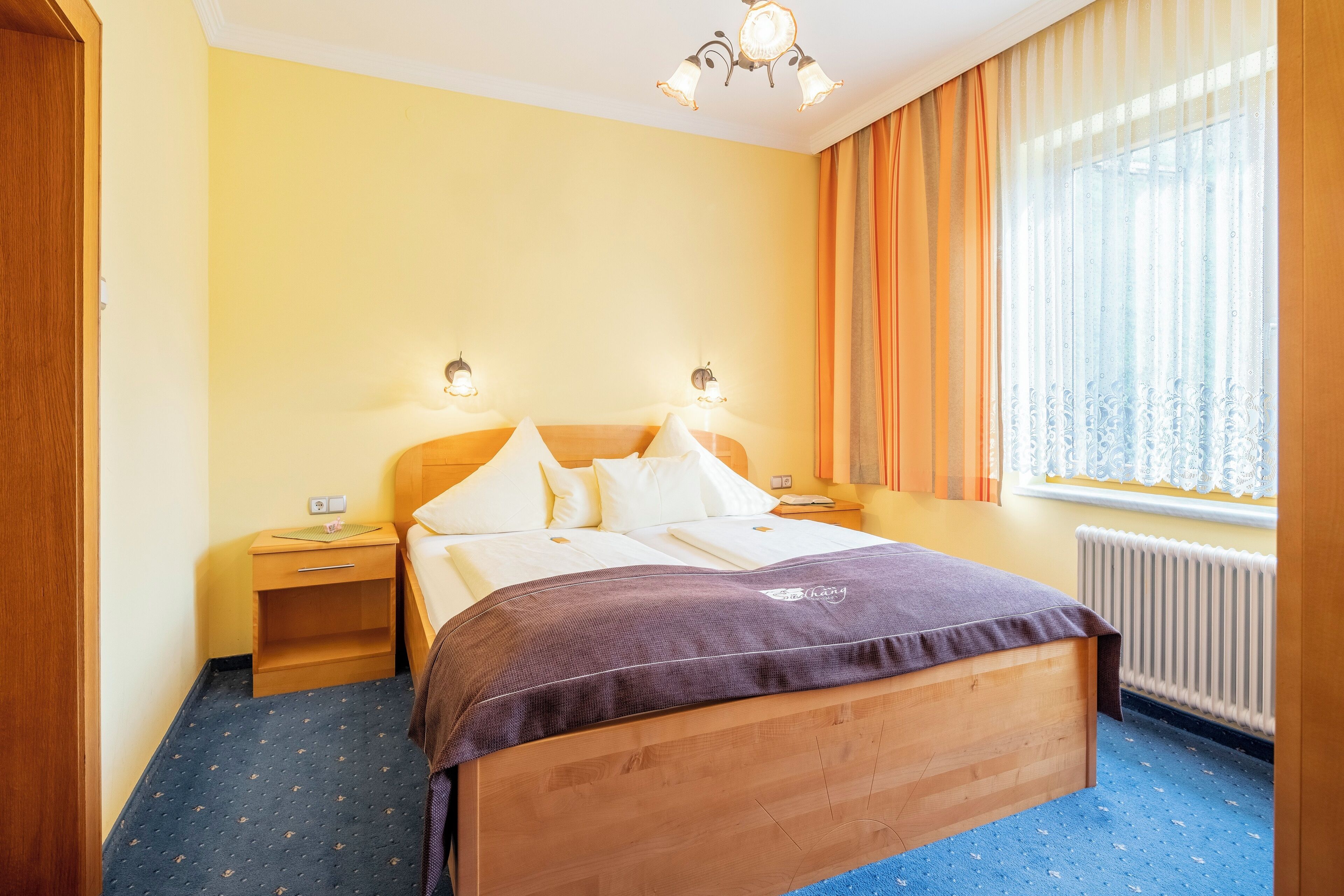 A hotel room with a wooden bed, two bedside tables, a nightstand, a window with curtains, and a ceiling light fixture.