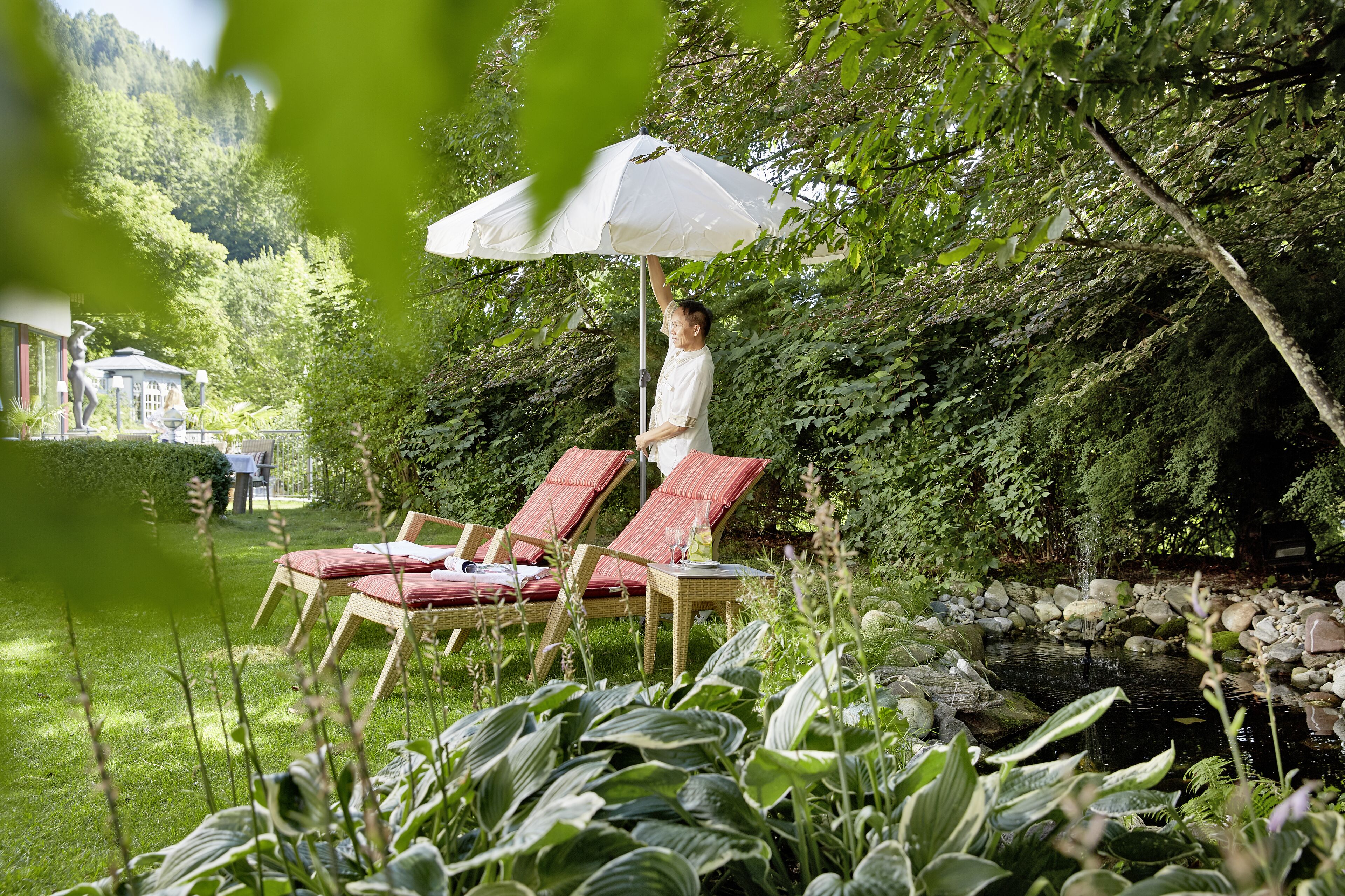 Un jardin avec un bassin, des fauteuils de jardin rouges et un parasol blanc.