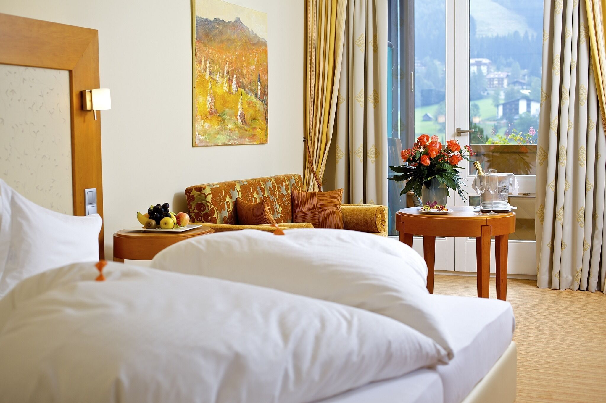 Habitación de hotel con una cama grande, un sofá, una pequeña mesa con frutero, un cuadro en la pared y una ventana con cortinas.