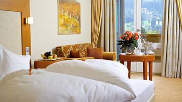 Habitación de hotel con una cama grande, un sofá, una pequeña mesa con frutero, un cuadro en la pared y una ventana con cortinas.