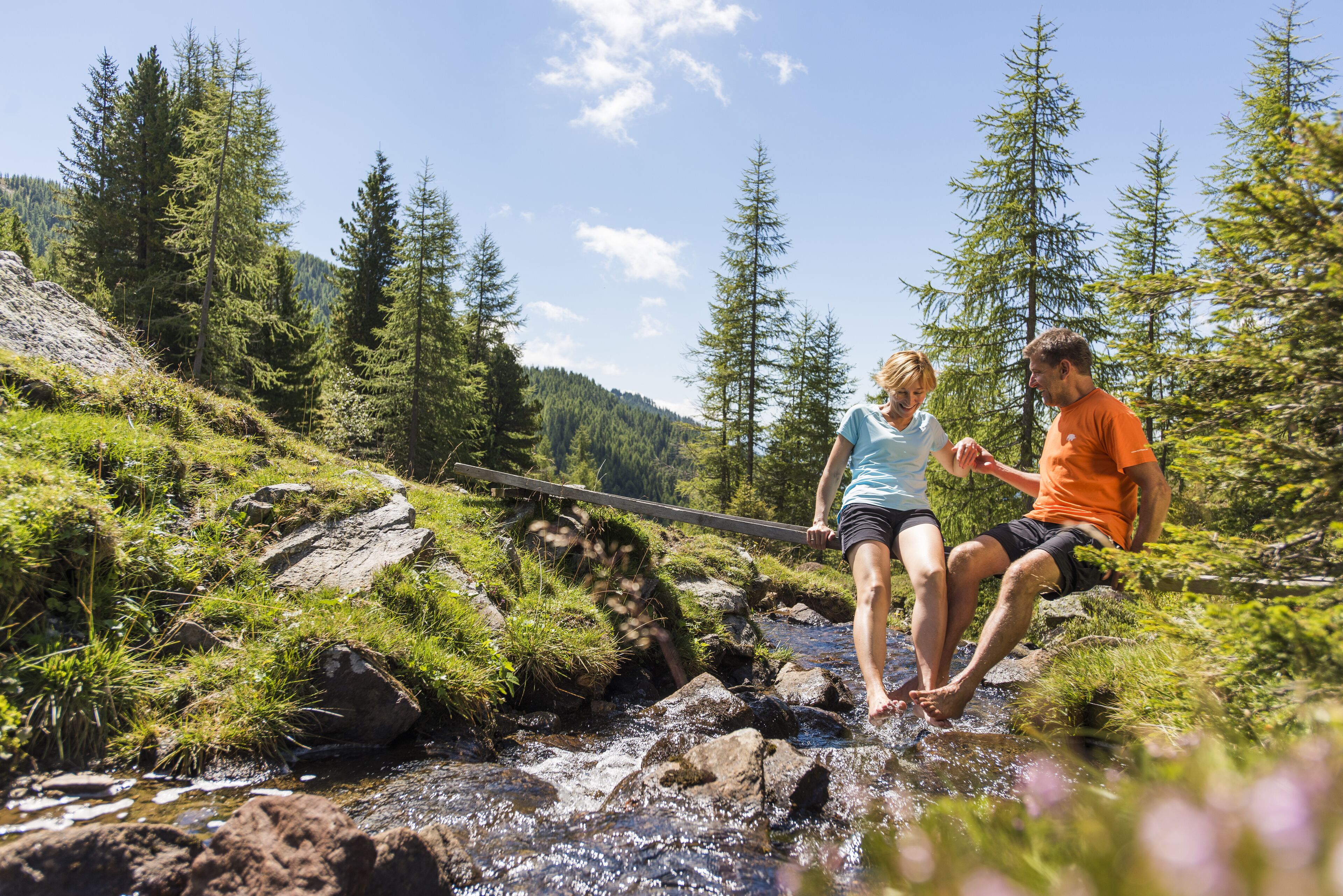 Two people standing by a stream in a forested area with trees and rocks.