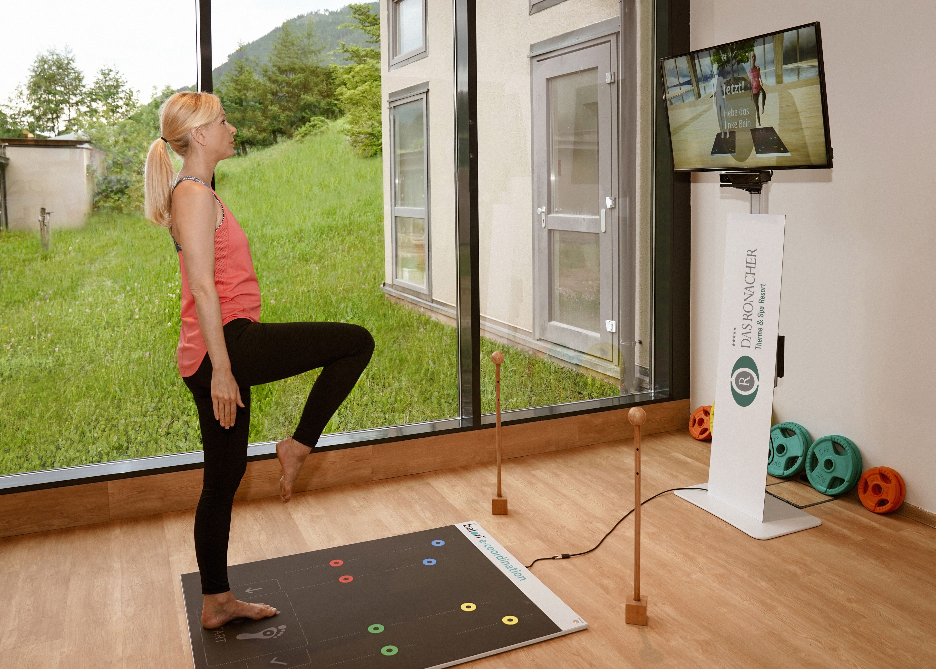 A yoga mat with a coordination exercise chart, a television displaying a yoga video, and a view of a green landscape through a window.