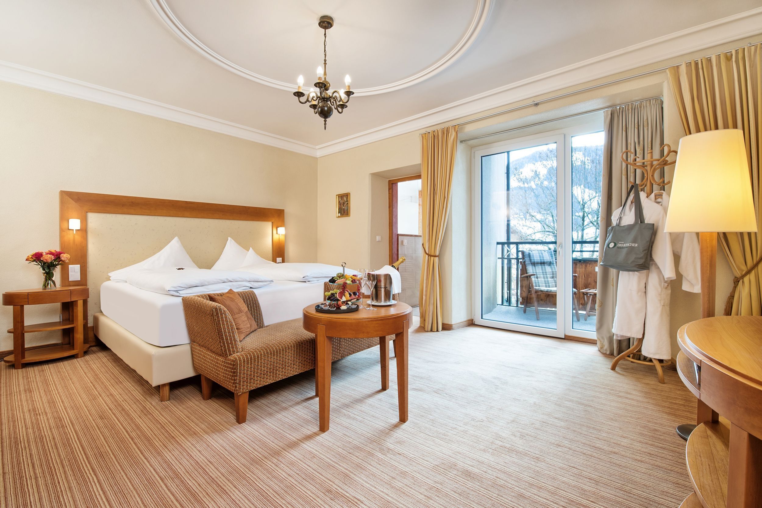 Une chambre d’hôtel avec un grand lit, une table de chevet en bois, une petite table ronde, une chaise, une lampe et une vue sur l’extérieur grâce à une porte-fenêtre coulissante.
