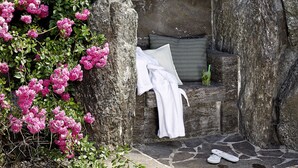 A stone bench with a white towel and a cushion, surrounded by large rocks and pink flowers.
